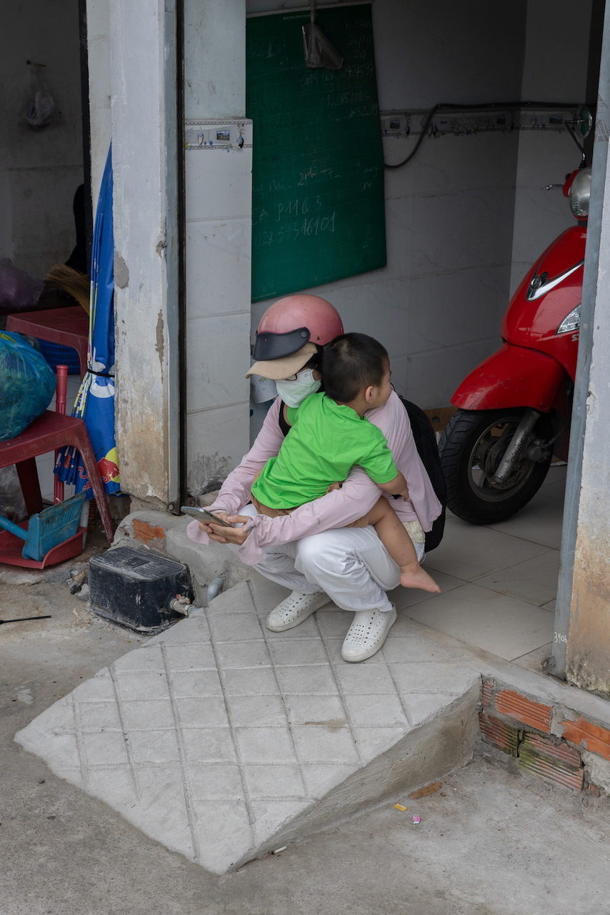 A woman crouches with a child on her back beside a red scooter.