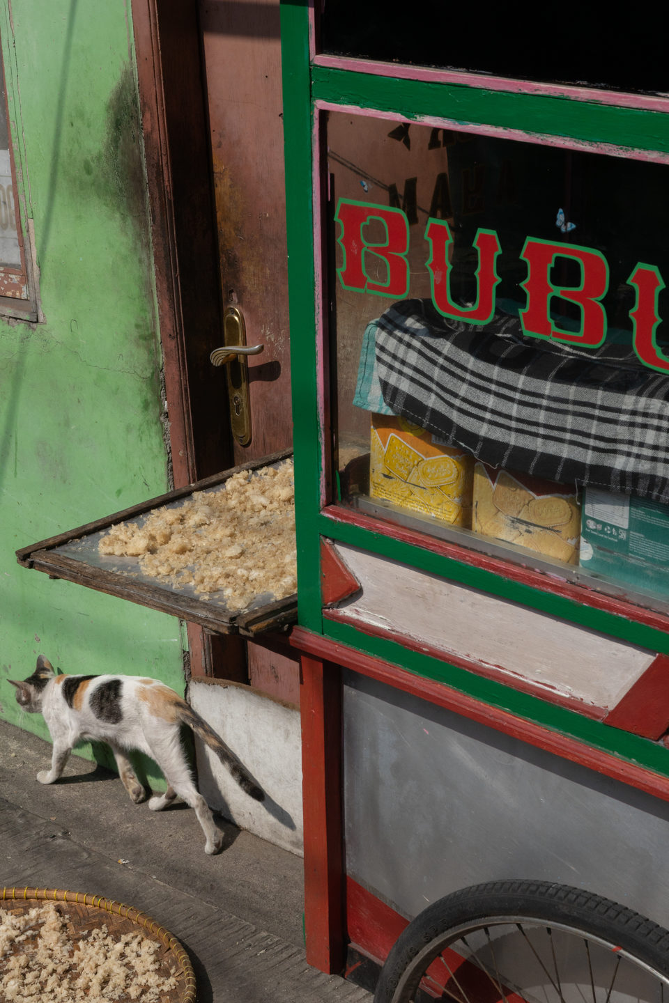 Red BUBUR lettering on a green cart, a calico cat crossing behind trays of shredded food.