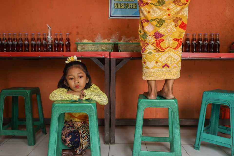 A girl rests on a green stool in a warung beside a woman in gold.