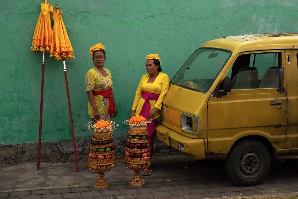 Two women in yellow with tiered offerings beside a yellow truck and green wall.