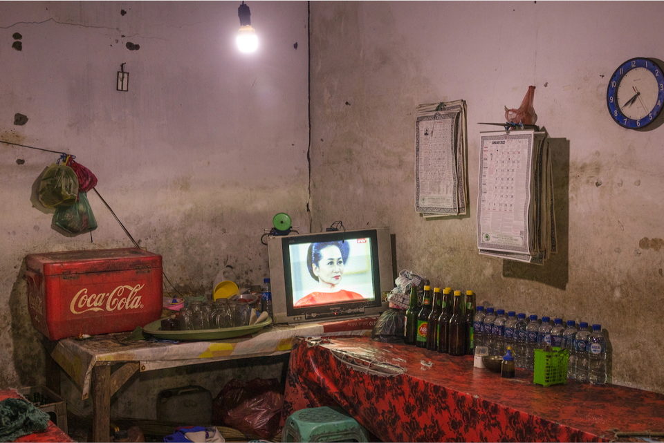 A warung interior in low light, a television glowing on the counter.