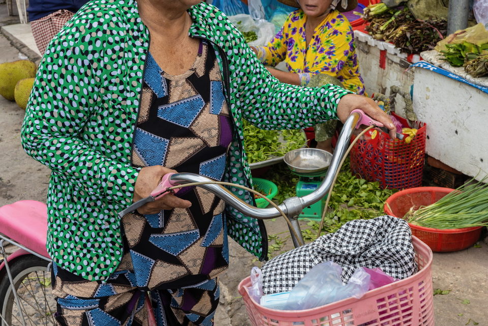 A woman in a patterned green jacket pushes a bicycle laden with market baskets.