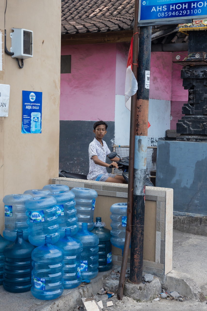A woman sits in a shopfront stacked with blue water bottles beside a red and white flag.