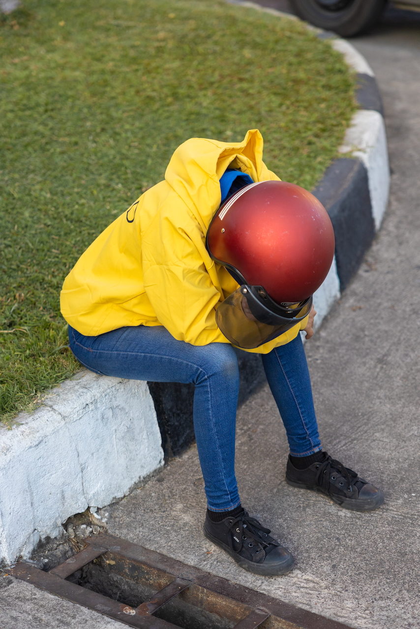 A person in a yellow jacket and red helmet rests head-down on a kerb beside cut grass.