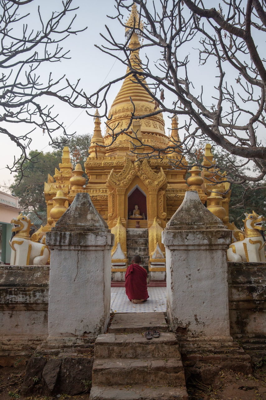 A monk in maroon robes walks between whitewashed gateposts toward a gilded pagoda.