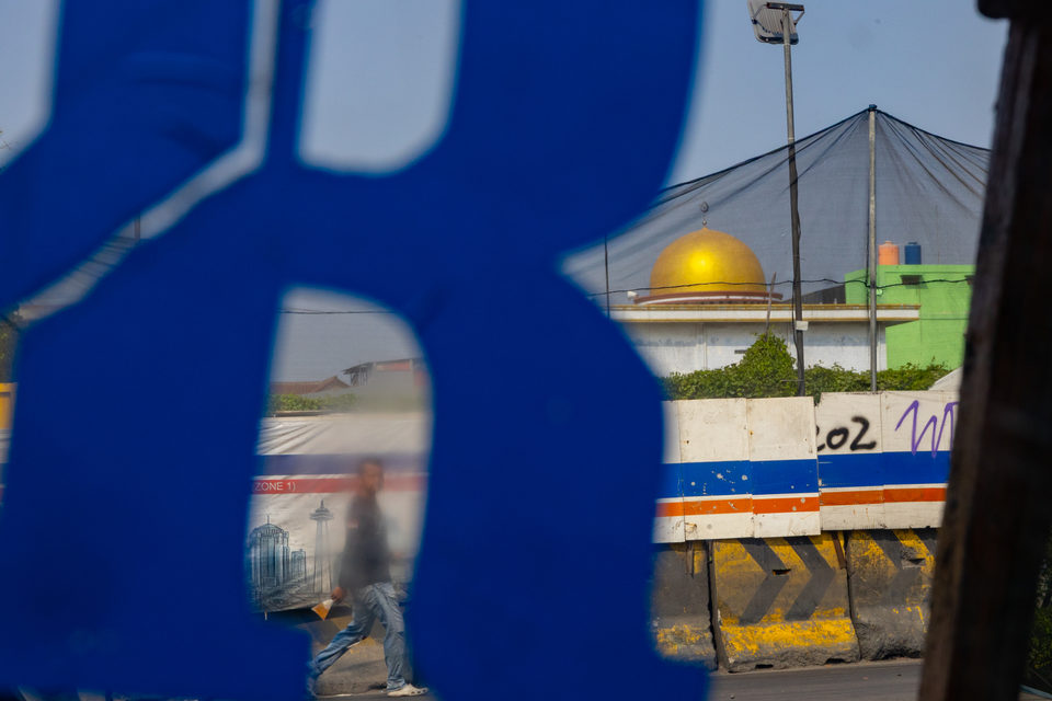 A blurred blue letter fills the frame, a gold mosque dome and a passing man behind.