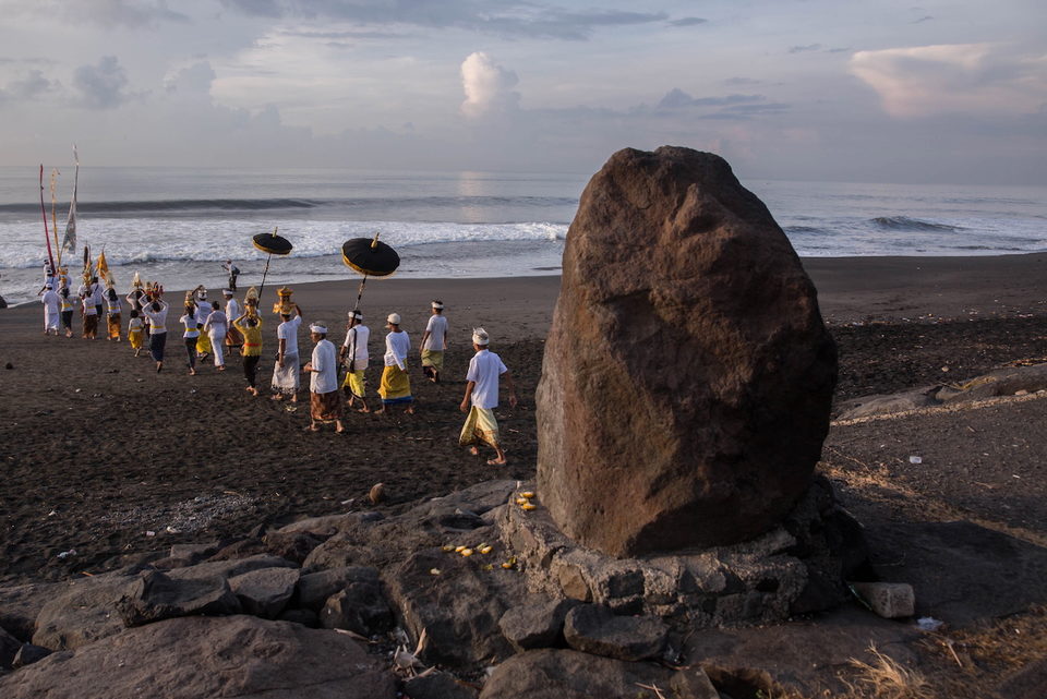 A procession walks a black sand beach past a large boulder.