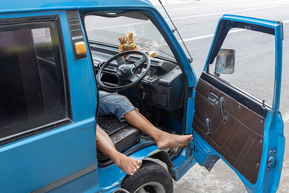 A man lies asleep across the seats of a blue truck, feet out the window.