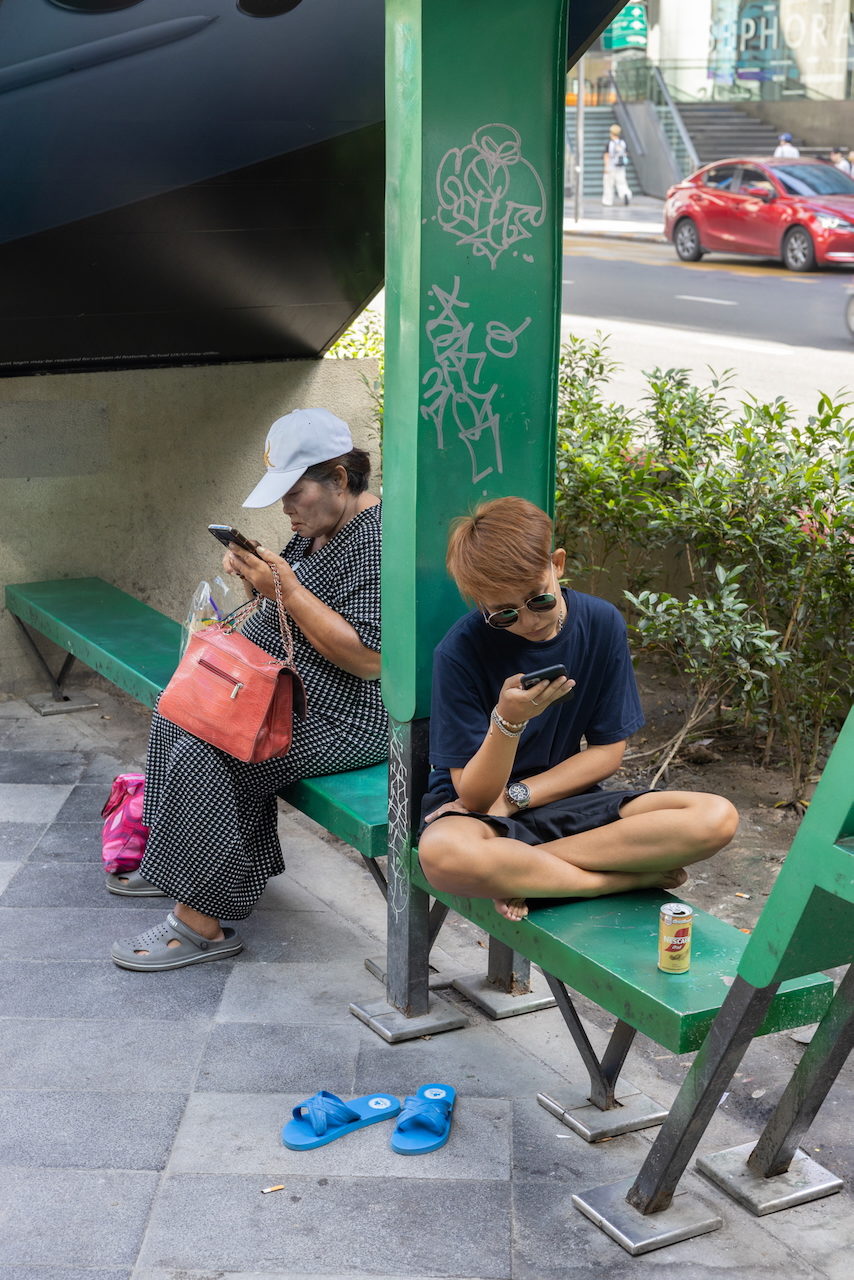 Two women sit on a green bench, both looking at their phones.