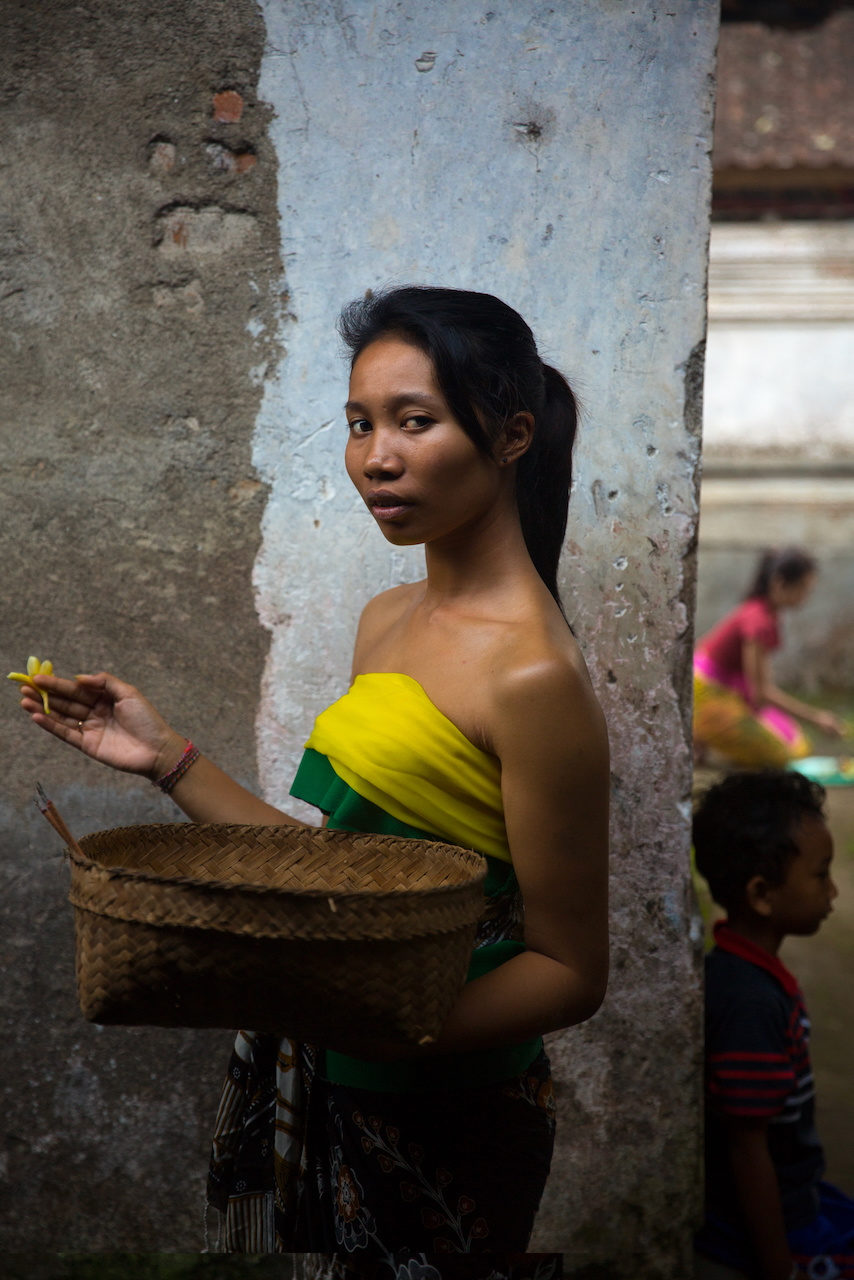 A young woman in a yellow and green sash holds a frangipani flower, a woven tray under her arm.