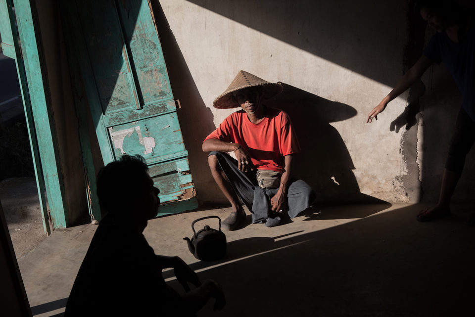 A man in a red shirt and conical hat sits in a shaft of hard sunlight beside a kettle.