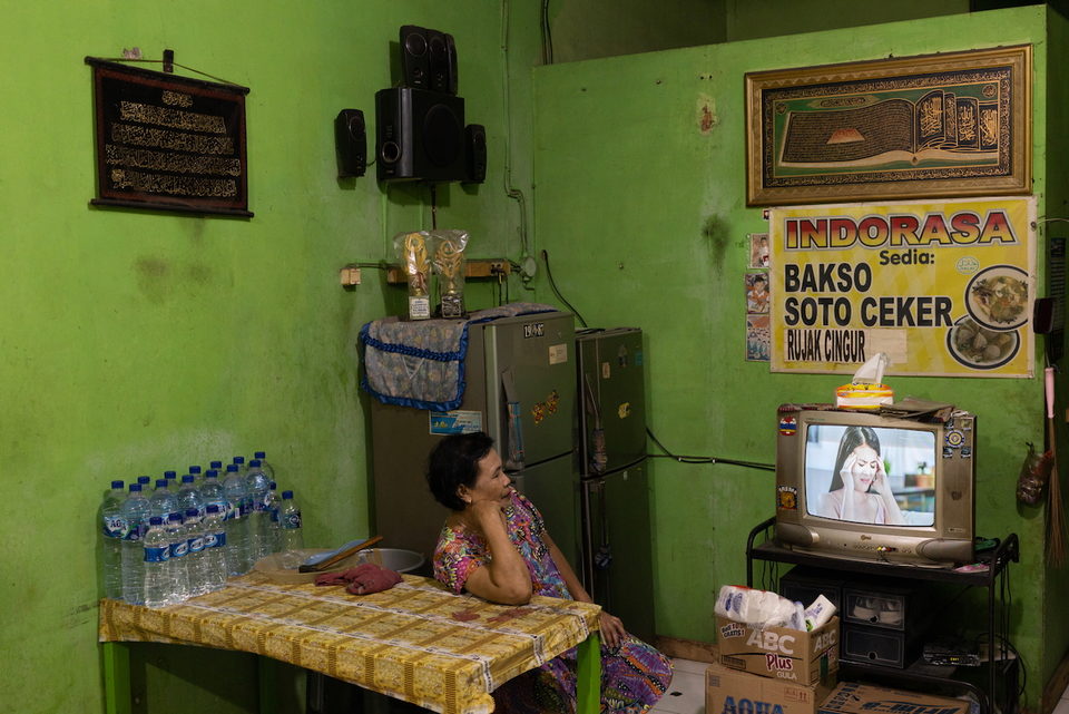A woman sits in a green-walled warung beside a fridge and a television.