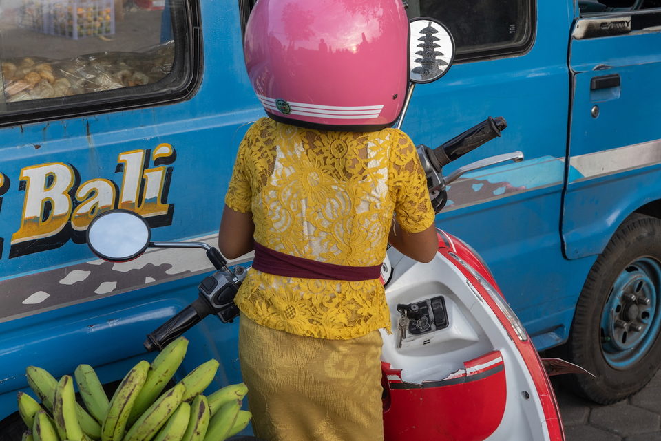 A woman in gold lace and a pink helmet stands by a scooter and bananas.