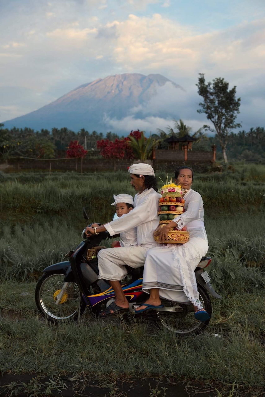 A family rides a motorbike carrying offerings, Mount Agung behind them.