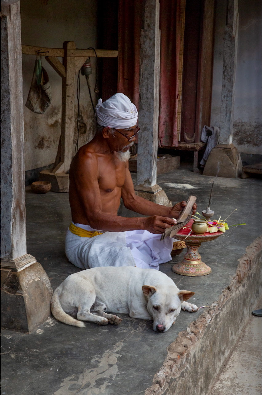 A bare-chested priest in a white headcloth prepares offerings beside a sleeping white dog.