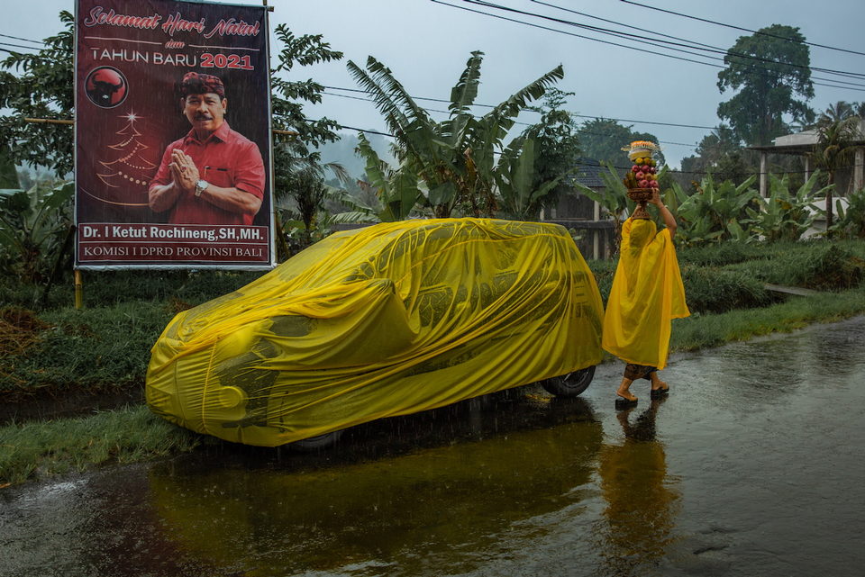 A car under a yellow cover in floodwater, a woman in a yellow poncho passing.