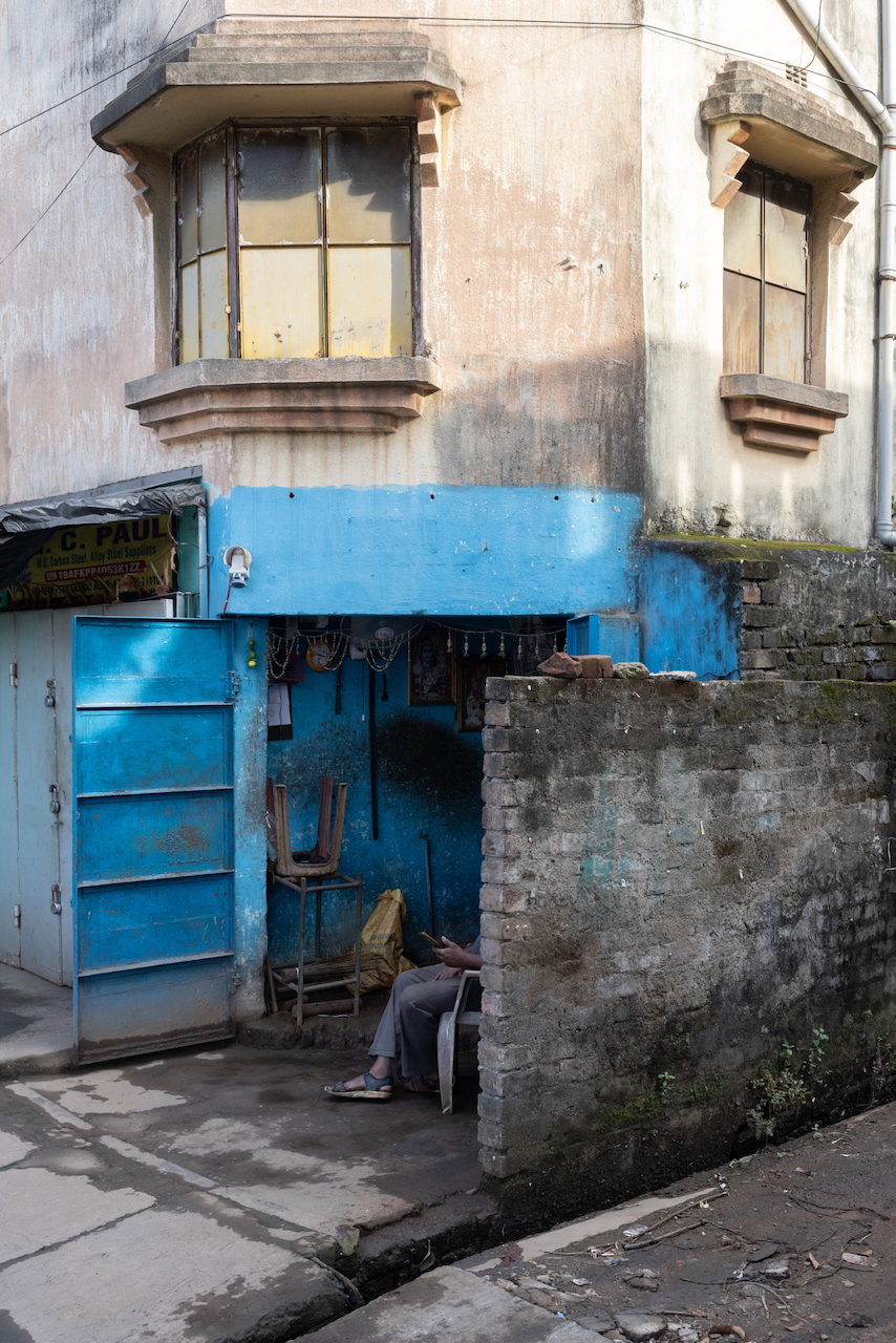 A blue-shuttered shopfront with a figure asleep in the shadow.