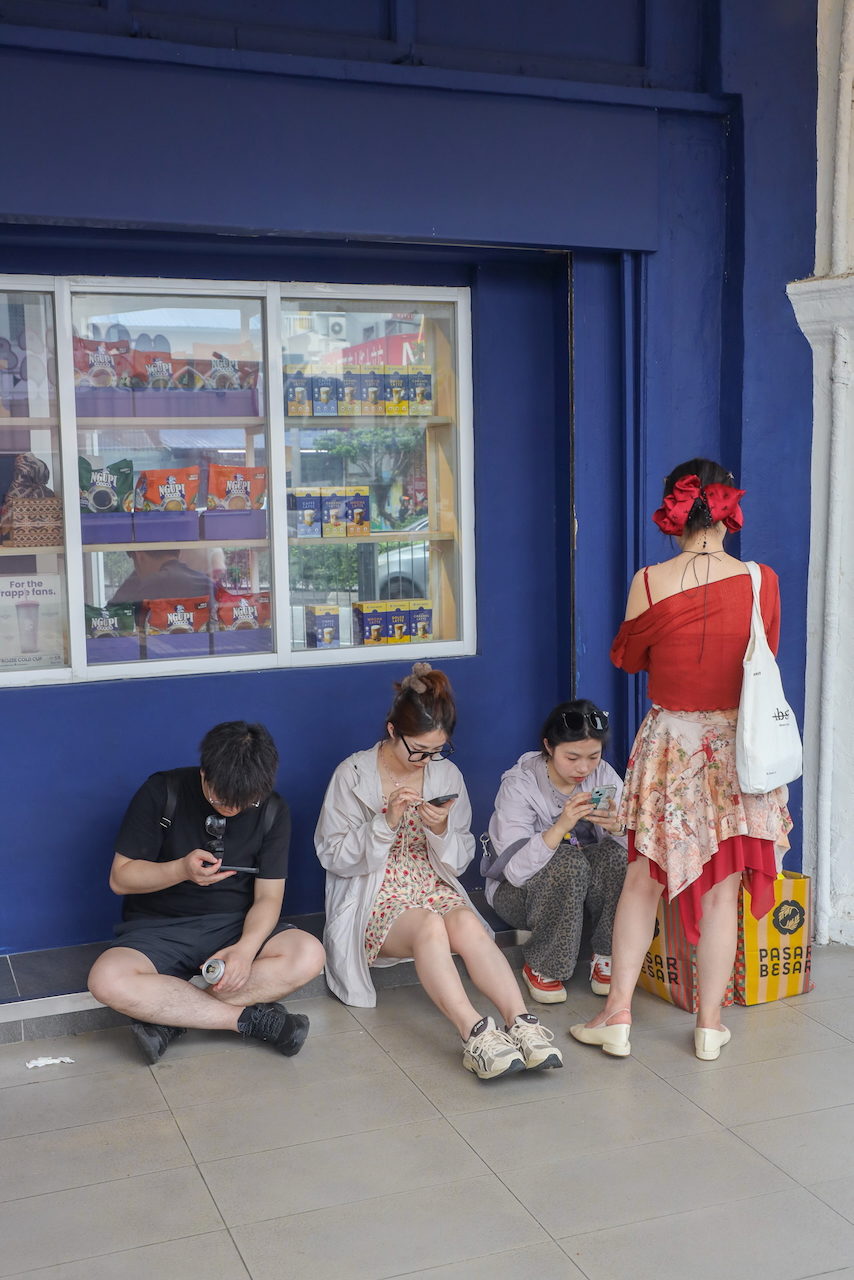 Young people sit against a blue shopfront, most looking at phones.