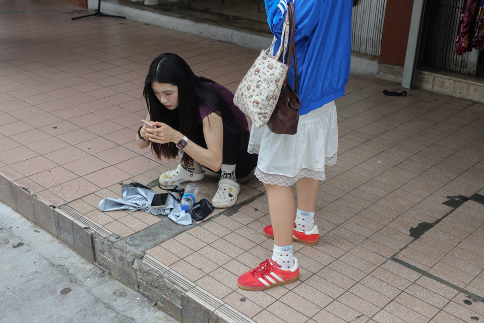 A woman crouches over shoes laid out on a footpath, phone in hand.
