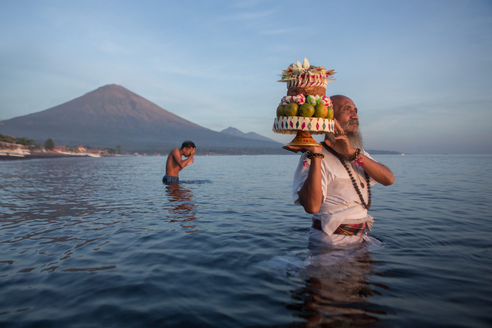 A man stands waist-deep in the sea carrying a tiered fruit offering, a volcano behind him.