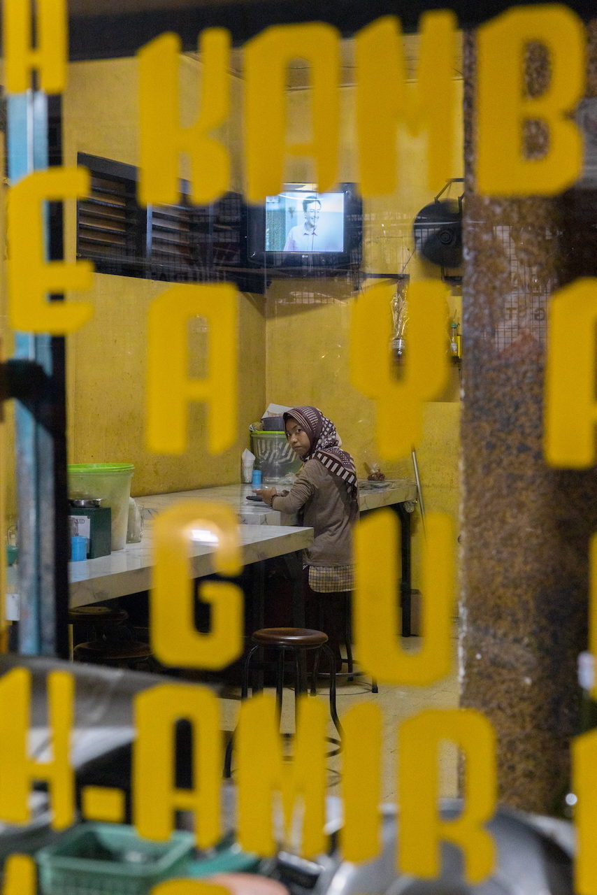 A woman sits in a warung behind yellow lettering on the window.