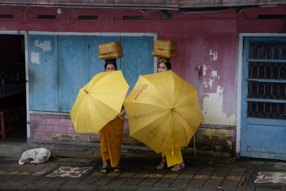 Two women shelter behind yellow umbrellas, offerings balanced on their heads, outside a blue and pink shopfront.