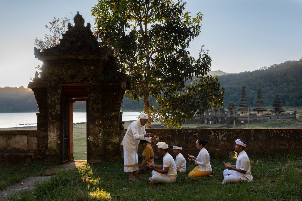 Worshippers kneel on grass before a lakeside temple gate.