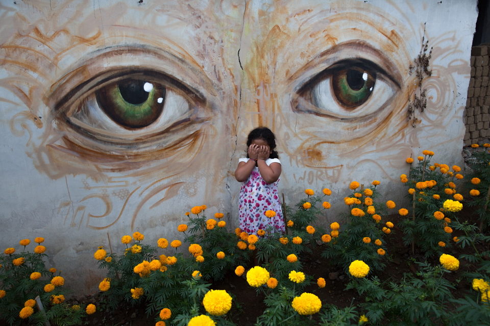 A small child stands among marigolds before a mural of large painted eyes.
