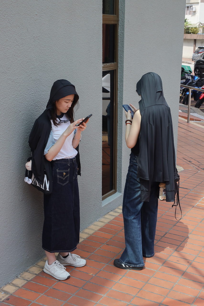 Two women in black stand against a wall, one looking at her phone.