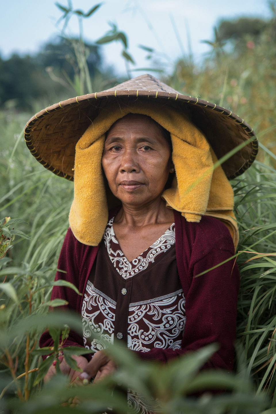 A woman in a conical hat and yellow headcloth stands in tall grass.