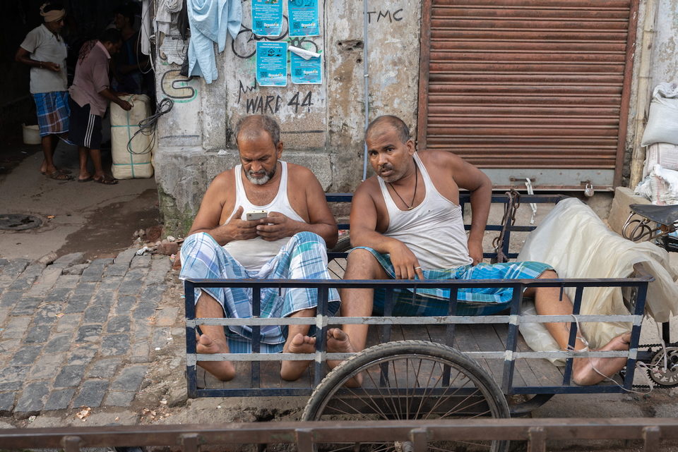 Two men in singlets sit on a cart, one looking at his phone.