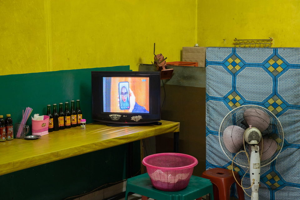 A television on a yellow counter beside bottles and a pink basin.