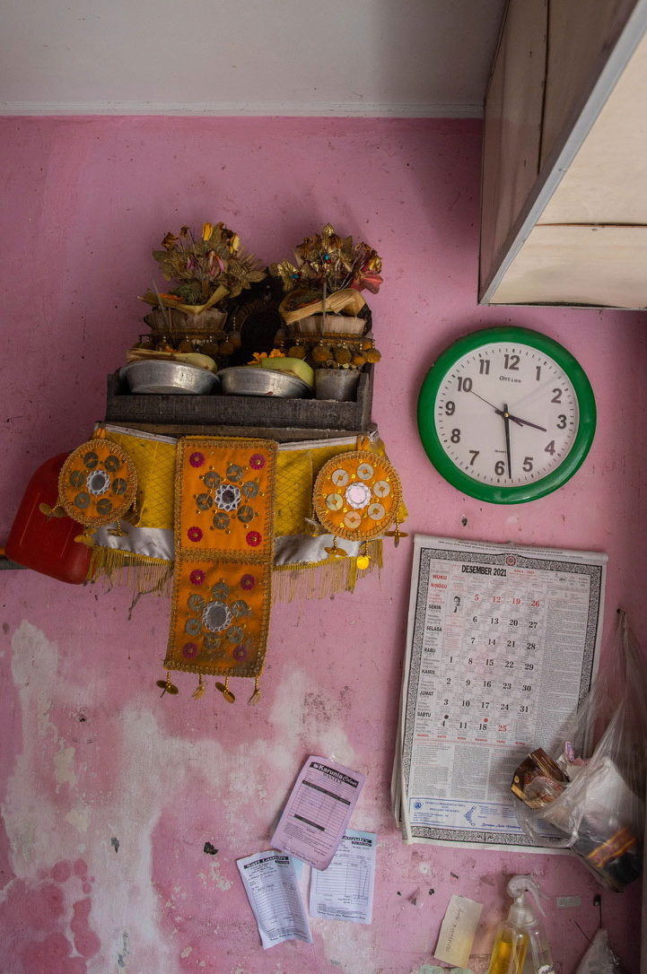 A household shrine with offerings and a green wall clock on a pink wall, beside a paper calendar.