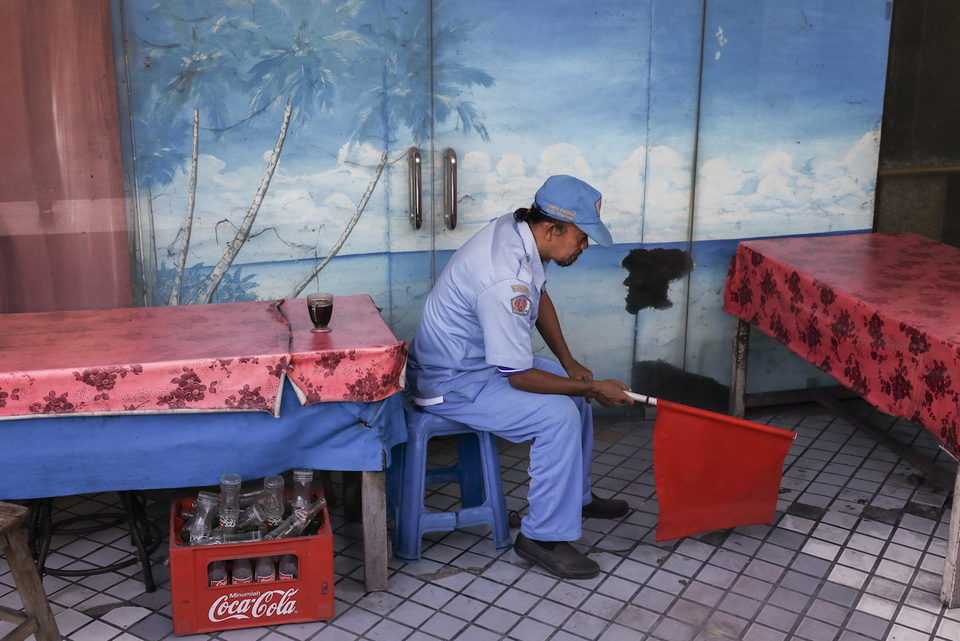 A man in a pale uniform and cap sits on a stool holding a red flag, against a painted seascape.