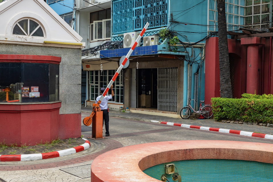 A person raises a red and white boom gate on a street of blue and white buildings.