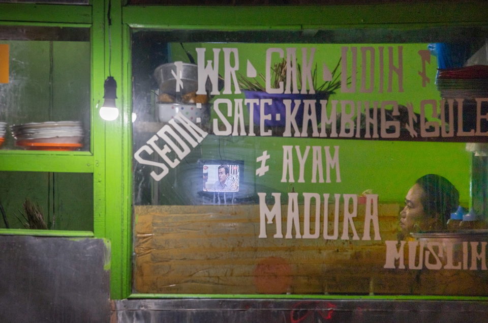 A hand-painted warung sign in green and white, a television behind the glass.