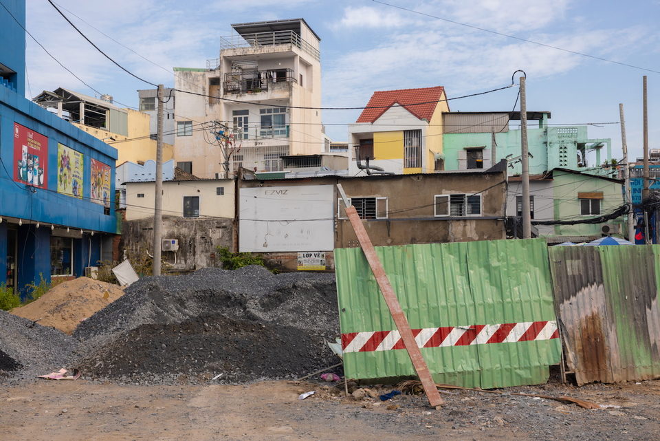 A green and red striped hoarding on a gravel lot in front of pale apartment blocks.