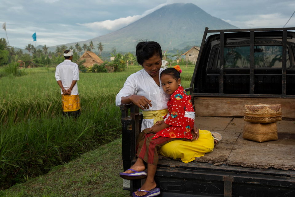 A family sits on the back of a truck in a green field, a volcano behind.