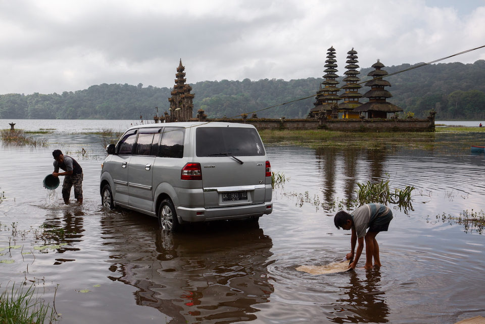 A silver van stands in shallow floodwater beside a lakeside temple with tiered meru roofs.