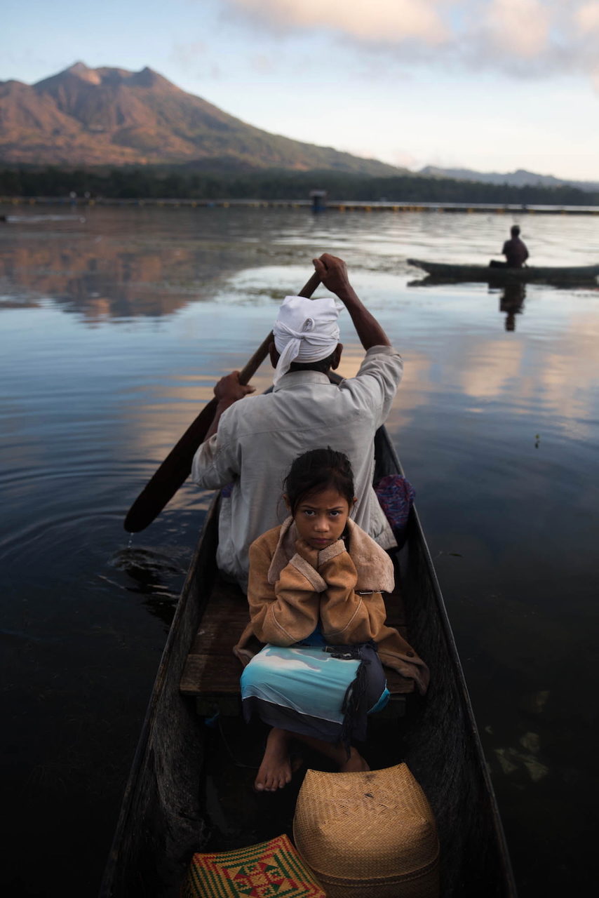 A man paddles a canoe with a child aboard on a still lake.