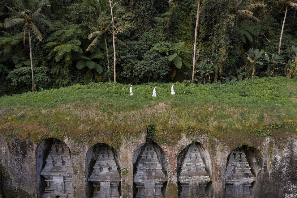 Small white-clad figures cross a green field above a row of stone shrines.