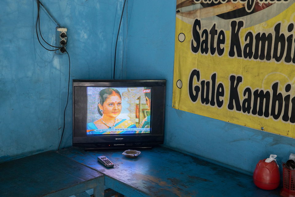 A television on a bench in a blue room below a satay banner.