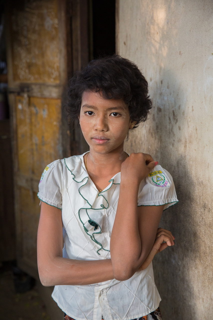 A young girl in a white embroidered blouse leans against a weathered wall, arms crossed.