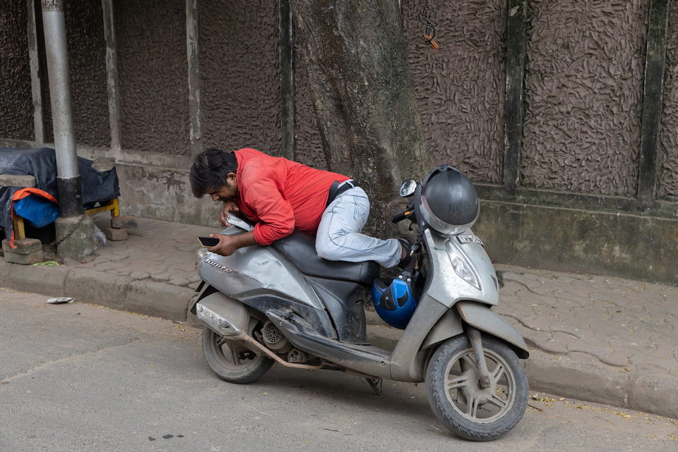 A man in a red jacket leans over his phone on a parked scooter.