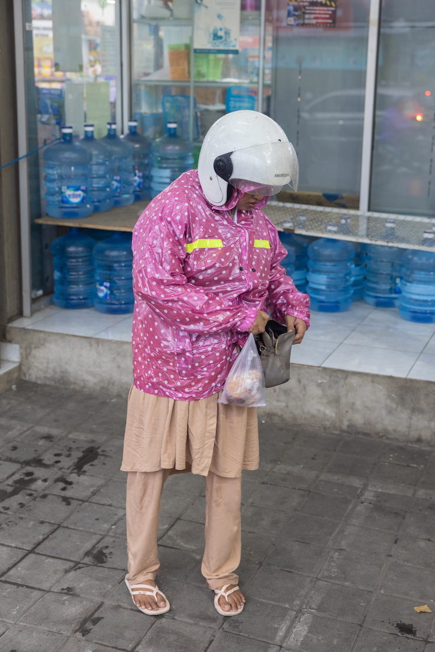 A woman in a pink patterned raincoat and white helmet stands before stacked water bottles.