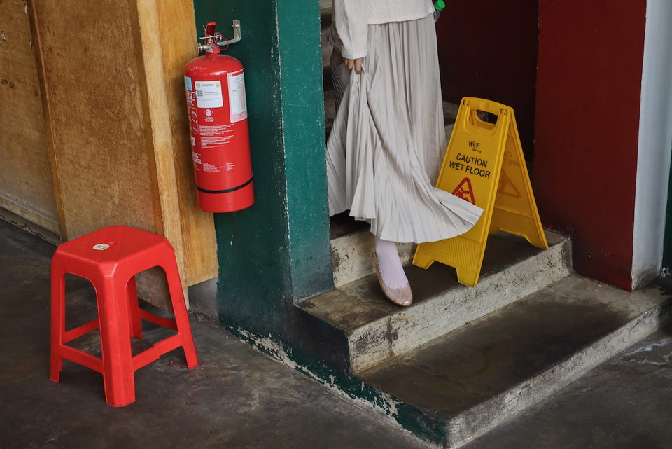 A figure in a pale robe steps down concrete stairs past a red fire extinguisher and a yellow wet-floor sign.