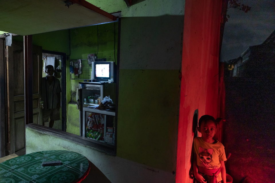 A child stands beside a red pillar, a television in the room behind.