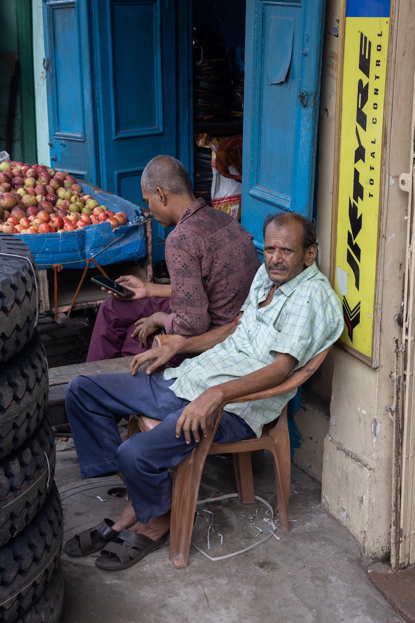 Men sit outside a fruit stall, one absorbed in his phone.