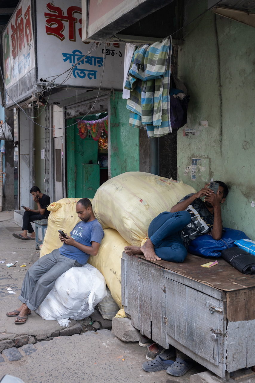 Men rest among sacks in a narrow street, one looking at his phone.