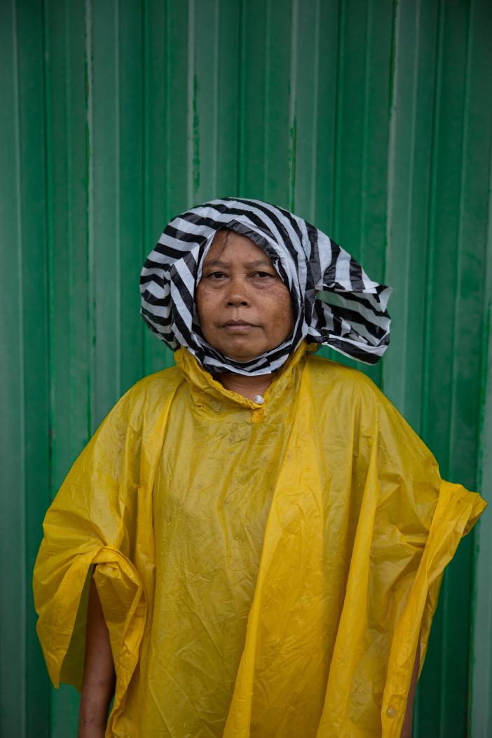 A woman in a yellow rain poncho and striped headscarf against a green wall.